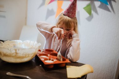 Parti şapkalı küçük bir çocuk, kek tepsisine kaşıkla vurmaya odaklanmış.. 