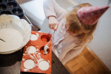 Genç bir kız küçük bir masada ev yapımı ikramlar hazırlıyor. Parti şapkası takıyor. Etrafı renkli süslemelerle çevrili..