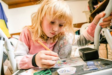 Sarışın kız evde elmas mozaik sanat eserleri yapmaya odaklanmış. Yoğunlaştırılmış ifade, dağınık bir odada siyah bir masanın üzerinde küçük mücevherler ve aletlerle el işi yapmak..