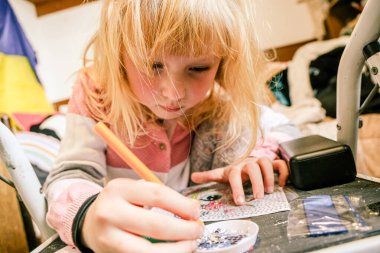 Sarışın kız evde elmas mozaik sanat eserleri yapmaya odaklanmış. Yoğunlaştırılmış ifade, dağınık bir odada siyah bir masanın üzerinde küçük mücevherler ve aletlerle el işi yapmak..