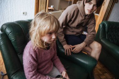 İki çocuk yeşil koltuklarda oturuyor, kendi düşüncelerine dalmışlar. Kız yumuşak bir kazak giyer ve erkek onu izlerken dalgın görünür. Güneş ışığı rahat odayı doldurur..