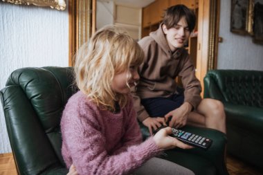 İki kardeş oturma odalarındaki yeşil koltuklarda rahatlar, birlikte televizyon izlerler. Küçük kardeş uzaktan kumandayı tutarken büyük kardeş onunla temas kuruyor ve sıcak bir atmosfer yaratıyor..