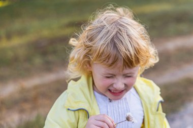 A small child with blonde curly hair smiles while holding a fluffy dandelion.