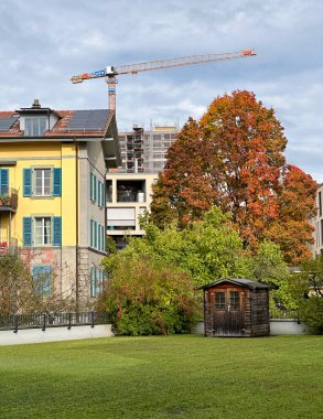 A yellow building with blue shutters and solar panels on the roof, located in Swiss urban neighborhood. The scene includes modern and traditional architecture with green foliage in the foreground, capturing the harmony between nature and urban life clipart