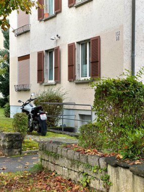 Kahverengi panjurlu bej bir bina ve girişte park edilmiş bir motosiklet. Bereketli yeşillik ve düşen sonbahar yaprakları bu sakin İsviçre mahallesindeki şehir manzarasına mevsimsel cazibe katıyor..