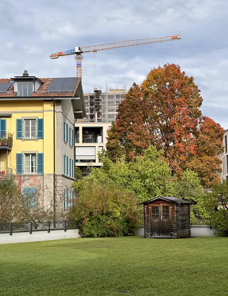 Çatısı mavi panjurlu ve güneş panelli sarı bir bina. İsviçre şehir merkezinde yer alıyor. Sahne, doğayla şehir yaşamı arasındaki uyumu yakalayan yeşil yapraklı modern ve geleneksel mimariyi içeriyor.
