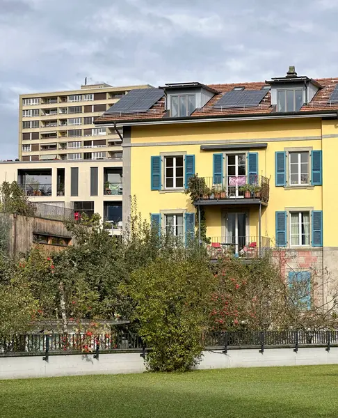 İsviçre şehir manzarasında mavi kepenkleri ve çatı güneş panelleri olan parlak sarı bir bina. Yeşillik ve modern yapılarla çevrelenmiş olan bina, gelenek ve sürdürülebilirliğin karışımını vurguluyor..