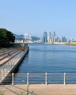 Busan 'da Gwangan Köprüsü, modern gökdelenler ve gezinti güvertesi boyunca oturma izlerinin yer aldığı sakin bir rıhtım sahnesi. Berrak mavi su ve uzak dağlar canlı şehir manzarasına derinlik katar..