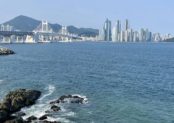 Gwangandaegyo Köprüsü 'nün suyun üzerinde uzandığı Busan şehri silueti. Arka planda yüksek binalar ve dağlar görünürken, ön planda kayalar ve deniz yer almaktadır..