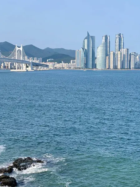 Busan şehrinin gökdelenleri ve Gwangan köprüsü engin mavi denizin üzerinde. Ön planda kayalık kıyı şeridi, uzak dağlar açık gökyüzünün altındaki şehir manzarasını çerçeveliyor..