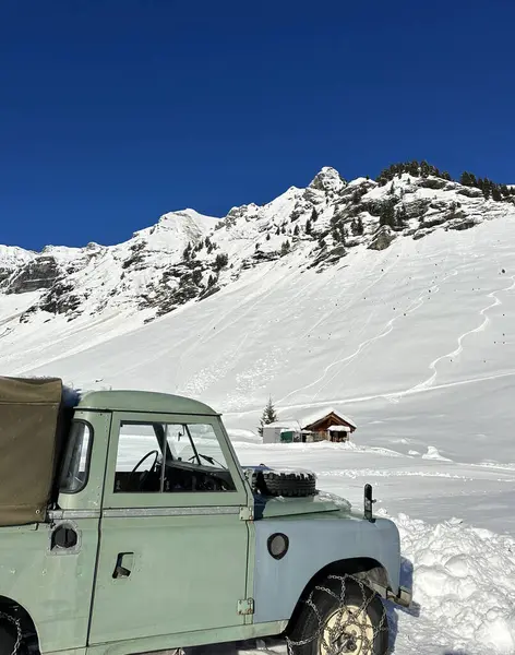 Lastiklerinde kar zinciri olan yeşil renkli bir off-road aracı İsviçre 'nin Murren kentindeki karlı bir arazide park halinde. Karla kaplı dağların zirveleri parlak mavi gökyüzünün altında yükselir. Arka planda bir ahşap dağ evi ve çam ağaçları görülebilir..