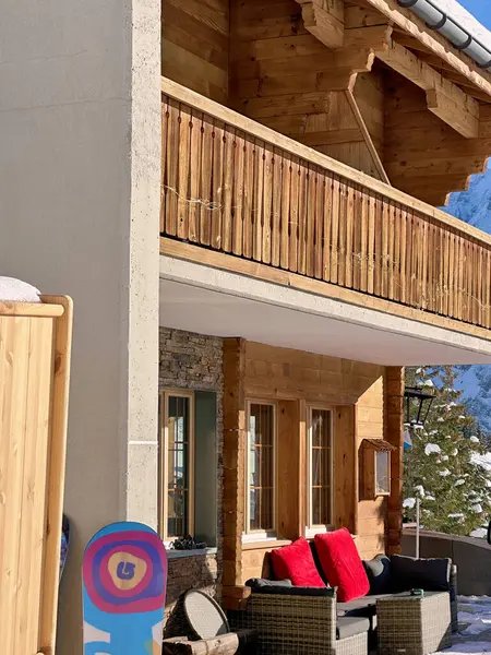 İsviçre, Mrren 'de ahşap bir dağ evinin terasında hasır koltuklarda kırmızı yastıklar var. Renkli bir snowboard duvara yaslanır ve arka planda parlak güneş ışığının altında karla kaplı bir çam ağacı görünür..