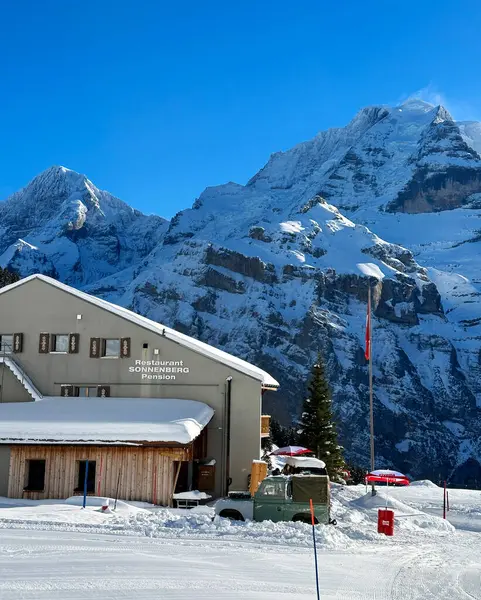 Restoran, İsviçre 'nin Murren şehrinde karla kaplı bir alana kurulmuş ve yüksek dağların nefes kesen manzarası var. Bir İsviçre bayrağı yeşil bir ağacın yanında dalgalanıyor. Parlak mavi gökyüzü kayak köyünün atmosferini yükseltiyor..