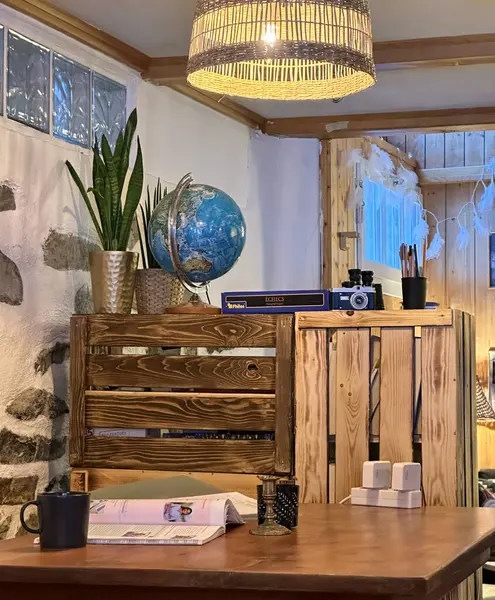 Açık dergi, siyah kupa ve mum tutacağı olan ahşap bir masa içeren sıcak, kırsal bir iç mekan. Bir küre, saksı bitkileri, ve ahşap bir rafta duran eski bir kamera, hasır bir abajur tarafından aydınlatılmış..