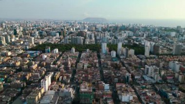 Lima şehrinin panoramik insansız hava aracı görüntüsü, merkezinde yemyeşil ve yapraklı bir park, arka planda ise heybetli bir uçurumun olduğu yerde, sakin bir yaz öğleden sonrasında,