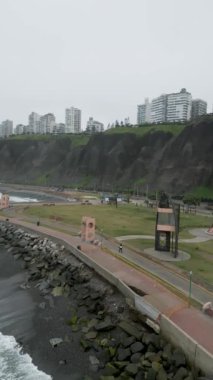 Peru 'nun Lima kıyılarında, parkta yürüyen insanların yanında sakin dalgalar ve trafiğin yoğun olduğu bir cadde ve arka planda bazı apartmanlar var.