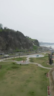 Peru 'nun liman kıyısındaki işlek bir caddenin yanındaki yeşil bir parkın hava manzarası bulutlu bir kış gününde alçak dalgalı sakin denizi görüyor.