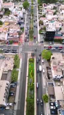 Araba trafiği ve kaldırımda yürüyen insanlar ağaçlarla çevrili bir caddenin kavşağında bir dron tarafından görüldüğü gibi binalar ve evlerle çevrili.