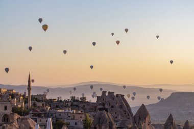 Arka planda görkemli dağların yükseldiği göz kamaştırıcı bir vadi manzarası, nefes kesici bir doğal manzara, şafak vakti sıcak hava balonları, Cappadocia 'daki volkanik tufa dağlarının ve mağara evlerinin zeminine karşı..