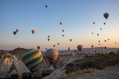 Arka planda görkemli dağların yükseldiği göz kamaştırıcı bir vadi manzarası, nefes kesici bir doğal manzara, şafak vakti sıcak hava balonları, Cappadocia 'daki volkanik tufa dağlarının ve mağara evlerinin zeminine karşı..