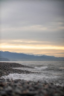 Arka planda görkemli dağların yükseldiği sakin bir plajın bulanık ve belirsiz bir resmi, pitoresk bir manzara yaratıyor.