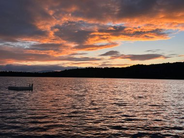 Tepe çizgili bir göl üzerindeki bulutlar yaz günbatımıyla turuncu renktedir. Bir yüzme botu göl sularının dalgaları arasında sessizce süzülür..