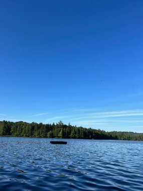 Huzurlu bir gölün dalgaları arasında yüzen bir iskele. Tepeler ve ağaçlar arka planda ve gökyüzü parlak mavi.
