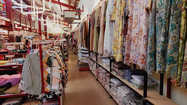 Pinrang-Endonezya, 28 Ekim 2024: Giysiler bir mağazadaki raflara asılıp düzgünce dizilmiş. Mağazanın koridorlarında çeşitli kumaş motifleri ve renkleri var..