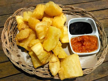 İçinde küçük kaplarda iki sos bulunan kızarmış kasava parçaları olan bir sepet tabağı: biri siyah sos ve biri kırmızı sos. Sepet ahşap bir masanın üzerinde..