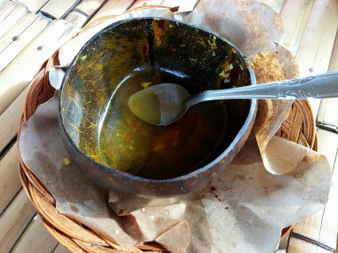 An empty coconut shell bowl with a spoon inside, placed on oil paper in a rattan basket. The bowl appears to have been used for soup. The background is a bamboo floor. clipart