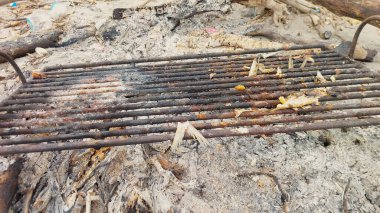 Üzerinde yanmış yiyecek olan kirli ve paslı demir ızgara. Bu ızgara kumlu zeminde, etrafı kül ve yanmış odunla kaplı..