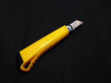 A cutter knife with a yellow plastic handle and a black tip, and a slightly visible metal blade. This tool is used for cutting paper, cardboard, or other thin materials.