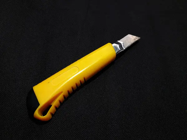A cutter knife with a yellow plastic handle and a black tip, and a slightly visible metal blade. This tool is used for cutting paper, cardboard, or other thin materials.