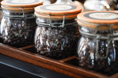 Kavrulmuş kahve çekirdekleri ile dolu cam kavanozlar, ahşap kapaklarla mühürlenmiş, ahşap bir tepsiye konmuş. Kavanozlar düzgünce düzenlenmiş ve odak noktası içindeki fasulyelerin dokusunu vurguluyor.