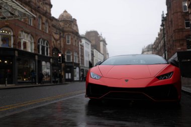 Yatay bir fotoğraf. Kırmızı bir spor araba tarihi İngiliz binalarıyla çevrili bir kaldırım taşı caddesine yakın park edilmiş. Şehir sabahın erken saatlerinde sisli..