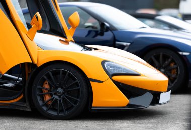 Kelebek kapılı parlak turuncu bir spor araba, bir kış günü İngiliz havaalanının otoparkında duruyor ve yanında mavi bir araba park ediyor. Arabanın ön kısmının yan görüntüsü..