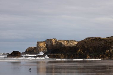 Sörf, okyanus ve kayalarla dolu bir kıyı şeridi. Yatay bir fotoğraf. Sörflü bir kıyı şeridi, sığ sularda dalgalar. Bir bahar günü arka planda çeşitli boyutlarda kayalar.
