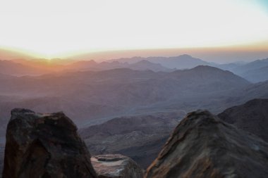 Güneşin doğuşundaki dağların manzarası. Ön planda, iki büyük kaya vadi ve dağ sırtlarının manzarasını oluşturur. Güneş ufukta görünüyor, gökyüzünü ve dağları turuncu ve pembe yumuşak tonlarda boyuyor..