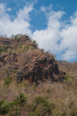 Mavi gökyüzü ve beyaz bulutlardan oluşan kuru ve çorak tepelerin manzarası. Kurak mevsim nedeniyle ağaçlar kurumaya başlıyor.