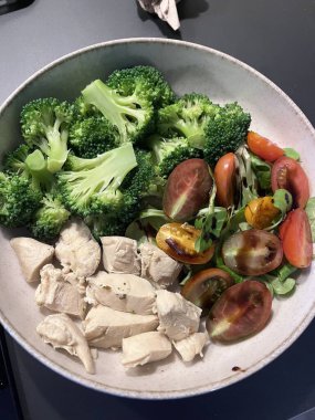 Bir kasede canlı brokoli ve renkli kiraz domateslerinin yanında pişirilmiş tavuk parçaları yer alıyor. Sağlıklı bileşenler ve dengeli beslenme içeren çekici bir yemek..