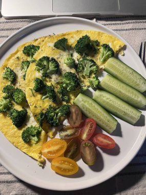 Brokoliyle doldurulmuş iki pofuduk omlet taze salatalık dilimleri ve renkli domateslerle birlikte bir tabakta servis edilir. Basit bir masada besleyici bir kahvaltı seçeneği..