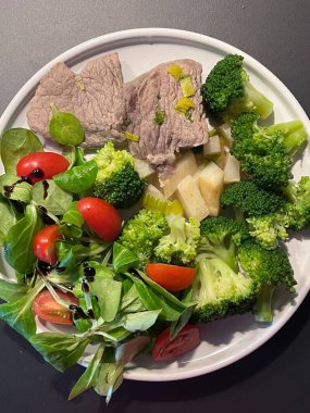 Brokoli, salata yeşillikleri ve vişneli domatesler de dahil olmak üzere çeşitli taze sebzelerin eşlik ettiği yumuşak et dilimleriyle dolu bir tabak. Bir balzamik jöle yağmuru tat katar..