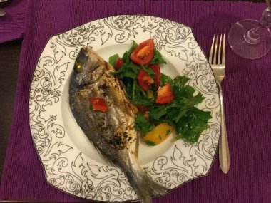 Güzel düzenlenmiş bir tabakta, kırmızı biberle süslenmiş ızgara bir balığın yanı sıra taze domates salatası ve yapraklı yeşillikler yer alıyor. Sunum renkli ve davetkar..