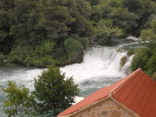 Güzel bir şelale, yeşil yaprakların arasında akar ve sakin bir atmosfer yaratır. Ön planda kırmızı fayanslı çatısı olan kırsal bir bina görülebiliyor ve manzaraya cazibe katıyor..