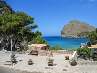 Sakin bir kıyı manzarası. Sakin mavi suların yanında kayalık bir uçurum. Yeşillik, taştan banklar ve canlı çömleklerle ilginç bir dinlenme alanı oluşturuyor..