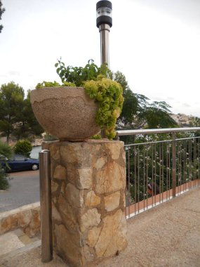 Canlı yeşil bitkilere sahip bir taş ekicisi taş bir kaidede sergilenir. Sahnede ağaçlar ve korkuluklar var. Yol kenarında huzurlu bir açık hava ortamı yaratıyorlar..