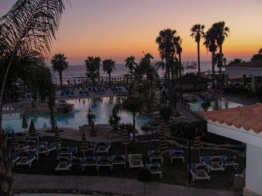 Titrek günbatımı, palmiye ağaçları ve sakin bir havuzu olan tropikal bir tatil köyünün üzerine güzel renkler salıyor. Gündüz geceye geçerken, misafirler huzurlu atmosferin tadını çıkarıyorlar..