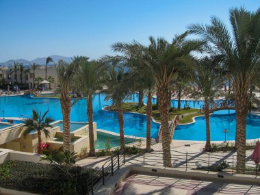 Berrak mavi bir gökyüzünün altında yemyeşil palmiye ağaçlarının arasına yerleştirilmiş birden fazla turkuaz havuzların yer aldığı sakin bir tatil köyü. Güneşli havanın keyfi ve rahatlığı için güneş pansiyonları etrafa saçılmıştır..