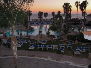 Sahil şeridindeki bir tatil köyünün üzerinden gökyüzünü saran huzurlu bir günbatımı. Palmiye ağaçları, güneşli bir havuzun etrafında şezlonglar ve plajda dinlenmek için sakin bir ortam yaratır..