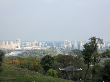Sonbaharda yemyeşil bir tepeden şehrin ufuk çizgisine bakan panoramik bir manzara. Sahne, sonbahar renklerini gösteren binalar, ağaçlar ve uzakta açık bir gökyüzü yakalıyor..